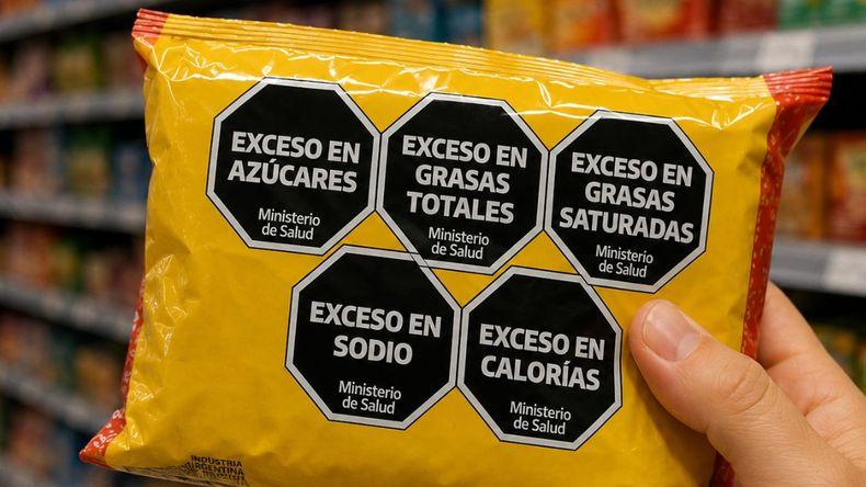 A packaged snack held on a supermarket aisle, showing black octagonal front-of-pack warning labels for excessive sugar, total fat, saturated fat, sodium and calories.